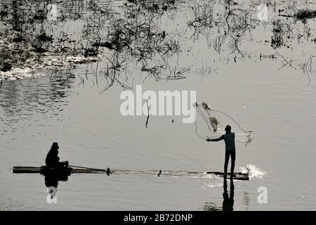 una silhouette di due uomini al sole della sera su una barca di bambù che cattura il pesce gettando una rete Foto Stock
