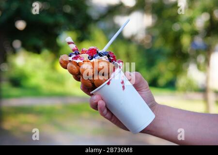 Waffle a bolle di frutta in tazza di carta bianca in mano su sfondo sfocato. Foto Stock