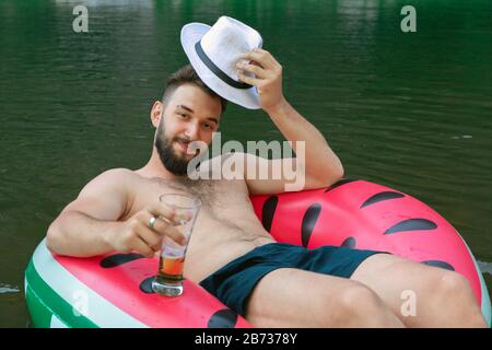 giovane attraente uomo sorridente toglie il cappello, accoglie. Giacente in anello di gomma con bicchiere di birra. Foto Stock