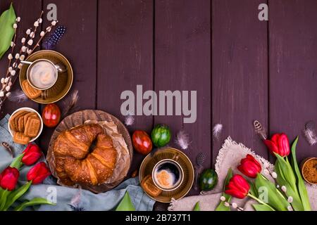 Colazione di Pasqua con caffè espresso fresco aromatico, croissant, torta brownie, uova colorate, tulipani rossi e salice. Caffè con dolci, fiori su un tavolo di legno. Vista dall'alto Foto Stock
