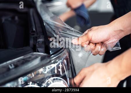 Processo di installazione PPF, stretching ravvicinata, rimozione manuale di una vecchia pellicola trasparente su un faro e cofano dell'auto. Servizio professionale automobilistico. Foto Stock