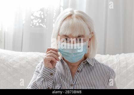 Donna bionda adulta in maschera medica siede sul divano in camera. Protezione da virus, coronavirus, ARVI Concept Foto Stock