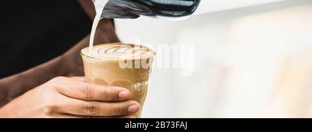 Striscione di barista professionista che versa il latte cotto a vapore in una tazza di vetro del caffè facendo bella arte latte Rosetta modello Foto Stock
