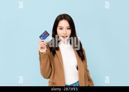 Bella donna asiatica sorridente in abiti invernali mostrando carta di credito in mano per i concetti finanziari e società senza contanti isolati sul retro azzurro Foto Stock