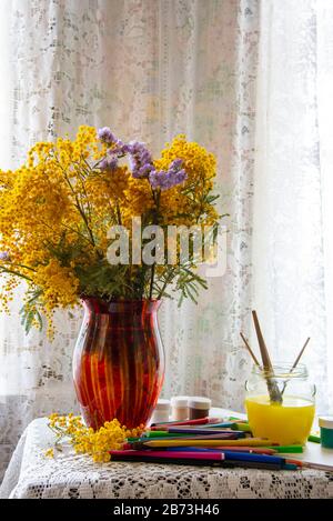 Matite colorate, pennarelli, e dipingere accanto ad un mazzo di Mimosa in un vaso rosso su una tovaglia di pizzo. Foto Stock