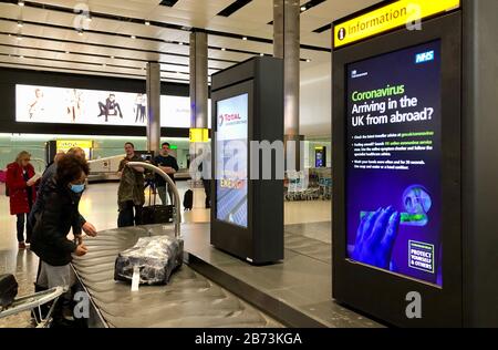 Avvertimenti del governo su Coronavirus nella Sala bagagli del Terminal 2 dell'aeroporto di Heathrow di Londra il giorno dopo che il primo ministro ha detto che Covid-19 "è la peggiore crisi di salute pubblica per una generazione", e il primo scienziato del governo ha avvertito che fino a 10,000 persone nel Regno Unito sono già infettate. Foto Stock