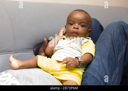 bambino neonato sdraiato sulle gambe del padre sul divano a casa guardando la macchina fotografica. Foto Stock