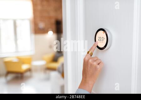 Donna che regola la temperatura di riscaldamento con un moderno termostato wireless installato sulla parete bianca a casa. Vista ritagliata messa a fuoco a portata di mano Foto Stock