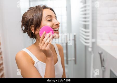 Giovane donna che pulisce la pelle del viso con speciale dispositivo a ultrasuoni durante le procedure di igiene mattutina in bagno. Cura della pelle e concetto di benessere Foto Stock