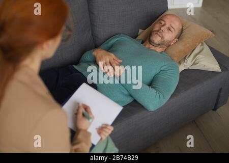 Uomo maturo sdraiato sul divano e che parla con il suo psicologo durante la sua visita in ufficio Foto Stock