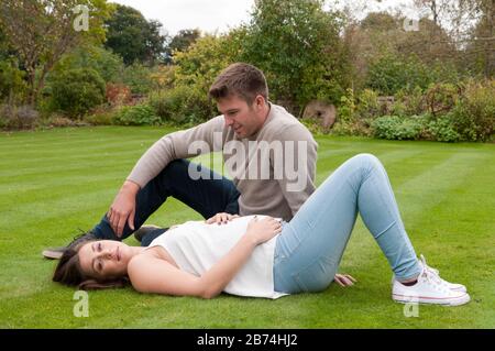 Bella donna incinta giovane sdraiata sull'erba con la sua partner seduto accanto a lei Foto Stock