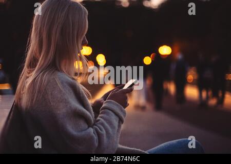 Giovane donna che usa lo smartphone mentre si siede sulla panchina in città al crepuscolo Foto Stock