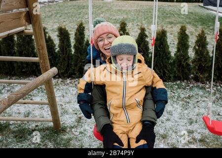 madre e figlio che giocano fuori ridendo mentre su un swing set Foto Stock