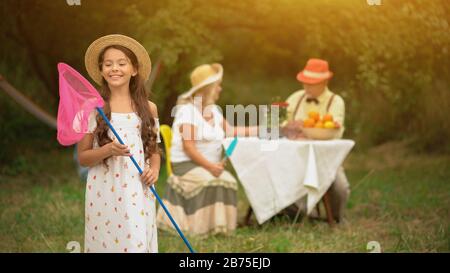 Ragazza giovane felice in UNA sundtress bianca e un cappello con una rete della farfalla nelle sue mani sta sorridendo e guardando la rete. I suoi nonni sono seduti al Th Foto Stock