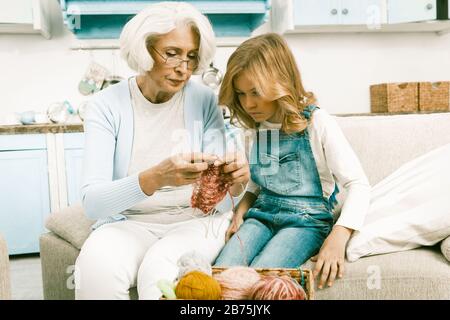 Nonna dai capelli grigi o bianchi che mostra la sua nipote come Knit mentre la sua visita seduta su divano o divano in soggiorno a casa su UNA cucina Ba Foto Stock