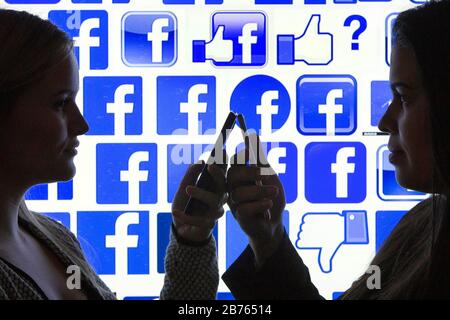 Le silhouette di due donne con gli smartphone in mano possono essere viste di fronte ai logo di Facebook, su 12.01.2016. Illustrazione agli amici di Facebook. [traduzione automatica] Foto Stock