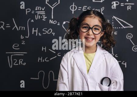 allievo sorridente della scienza della ragazza con gli occhiali in cappotto di laboratorio sullo sfondo della lavagna della scuola con disegno della mano modello di formula della scienza, di nuovo a schoo Foto Stock
