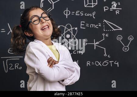 divertente bambina studentessa di scienza con occhiali in lab coat ridere su sfondo lavagna scuola con disegni a mano scienza formula modello, torna a s. Foto Stock
