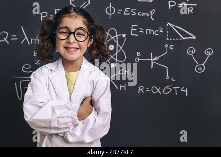 allievo sorridente della scienza della ragazza con gli occhiali in cappotto di laboratorio sullo sfondo della lavagna della scuola con disegno della mano modello di formula della scienza, di nuovo a schoo Foto Stock