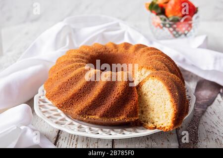 Un tavolo di legno bianco con una torta di bundt, kugelhupf o sockerkaka. Con una ciotola di fragole, un tovagliolo bianco e una taglierina per torte d'annata nel backgro Foto Stock