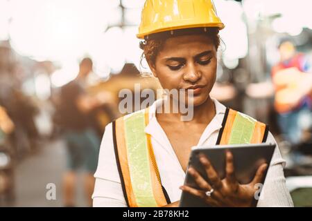 Donne americane lavoratrici in pesante industria manutenzione ingegnere felice di lavorare indossando uniforme di sicurezza e casco utilizzando tablet computer per controllare la macchina Foto Stock