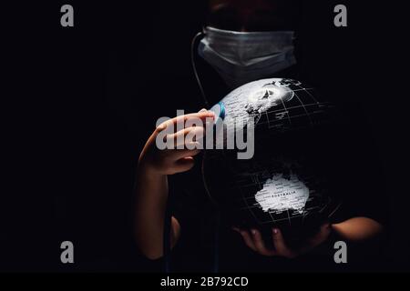 Mani di bambino tenere globo mettere stetoscopio sulla sfera ascoltare heartbeat, faccia coperta in maschera su sfondo nero. Problemi ecologici disastri e COVID- Foto Stock