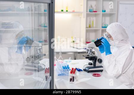 Giovane scienziato o ricercatore contemporaneo in tute protettive, maschera e guanti che cercano al microscopio mentre studiano nuovo virus Foto Stock