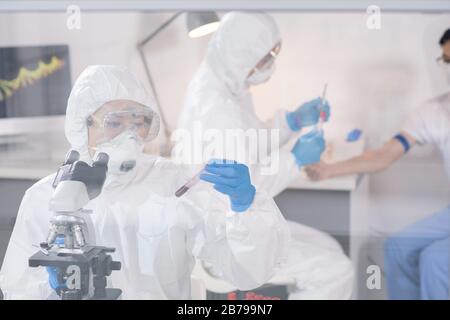 Giovane ricercatrice femminile in tute protettive che guarda fiasca con campione di sangue mentre studia virus durante il lavoro sul nuovo vaccino Foto Stock