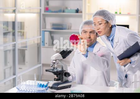 Giovane farmacista femminile e la sua collega che guardano la nuova sostanza chimica nella piastra Petri mentre studiano le sue caratteristiche Foto Stock