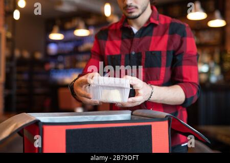 Giovane corriere di caffè che mette il contenitore con il cibo in grande sacchetto mentre imballano gli ordini dei clienti prima di consegnarli Foto Stock