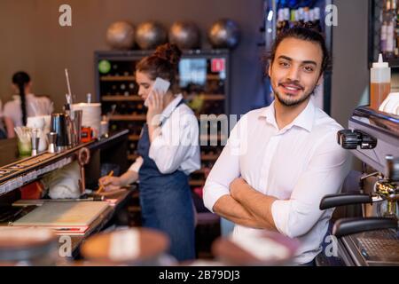 Cameriere giovane e allegro con armi incrociate in camicia bianca davanti alla telecamera sullo sfondo del collega che parla al telefono con il cliente Foto Stock