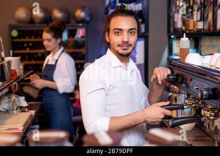 Giovane cameriere in camicia bianca che prepara cappuccino fresco con macchina da caffè mentre il suo collega puliva il vetro sul posto di lavoro Foto Stock