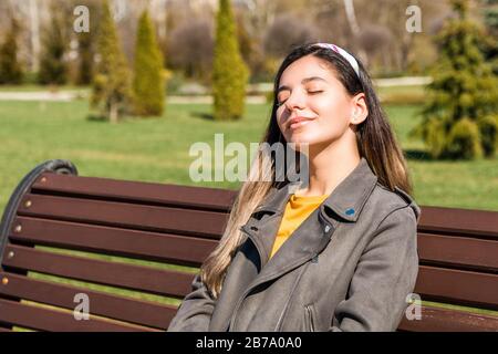 bella giovane hipster donna seduta su panchina in parco con gli occhi chiusi godendo di raggi di sole primavera Foto Stock