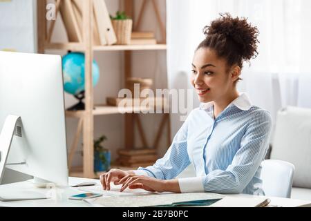 Discesa africana donna tour agente seduto al tavolo in ufficio digitando le informazioni di prenotazione sul computer sorridente allegro concentrato Foto Stock