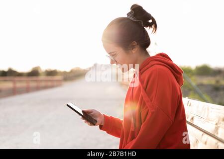 Giovane donna con gli occhiali sui suoi venti anni che chiacchierano sul suo telefono mentre siede in una panchina di un parco. Sorride e indossa un maglione rosso e un pony Foto Stock