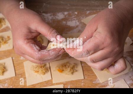 Una donna scolpisce gnocchi e ravioli da quadrati di pasta e cavolo. Pannello di taglio compensato, setaccio di farina di legno e perno di laminazione di legno - attrezzi per Foto Stock