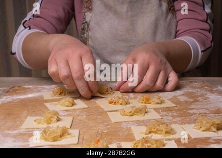 Una donna scolpisce gnocchi e ravioli da quadrati di pasta e cavolo. Pannello di taglio compensato, setaccio di farina di legno e perno di laminazione di legno - attrezzi per Foto Stock