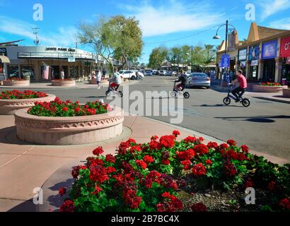 Tre giovani su scooter elettrici velocità vicino gerani rossi in primo piano e gallerie, ristoranti e quartiere dello shopping a Scottsdale, Arizona Foto Stock