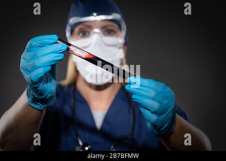 L'operatrice da laboratorio che indossa la maschera medica per il viso trattiene la provetta di sangue contro lo sfondo scuro. Foto Stock