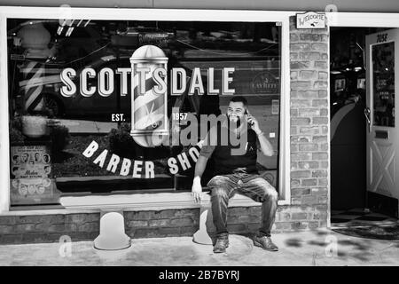 Un uomo sorridente, barbuto, alla moda sul telefono cellulare si siede sul banco di fronte allo Scottsdale Barber Shop, nella città vecchia, Scottsdale, Arizona, Stati Uniti, in nero e whit Foto Stock