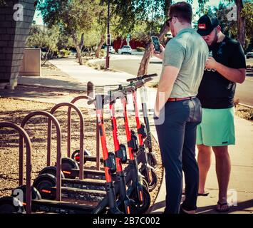 Due uomini interagiscono sui loro telefoni cellulari per navigare il noleggio di uno scooter elettrico, in rack vicino marciapiede nella città vecchia Scottsdale, Arizona, Stati Uniti Foto Stock