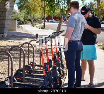 Due uomini interagiscono sui loro telefoni cellulari per navigare il noleggio di uno scooter elettrico, in rack vicino marciapiede nella città vecchia Scottsdale, Arizona, Stati Uniti Foto Stock