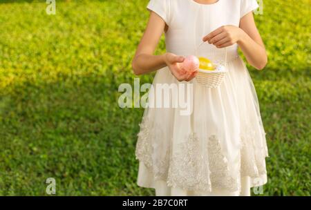 Ragazza carina che tiene uova di Pasqua colorate in un cestino. Bambino che si diverte all'aperto. Bambino che gioca con le uova sull'erba verde. Concetto di vacanze di primavera. Foto Stock