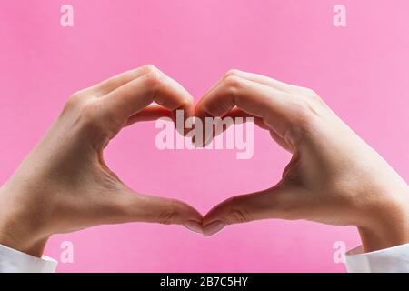 Mani femminili che formano un simbolo del cuore su sfondo rosa Foto Stock