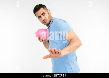 giovane uomo in una t-shirt blu tiene una banca piggy e disaccetta mostrando il gesto di rifiuto su uno sfondo bianco studio Foto Stock