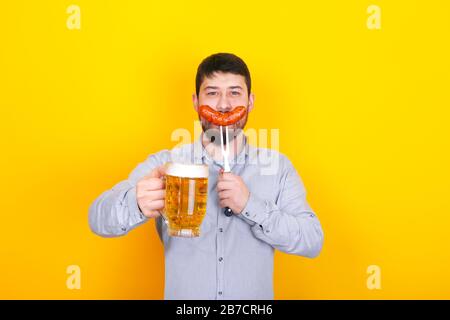 uomo con un bicchiere di birra e salsiccia alla griglia su una forchetta in mano, in piedi su sfondo giallo Foto Stock