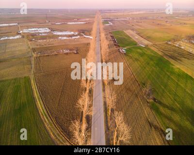 Golden hour paesaggio aereo di una strada circondata da alberi e praterie con alcune macchie di neve lasciato su di loro a Mihail Kogalniceanu, Ialomita, Rom Foto Stock