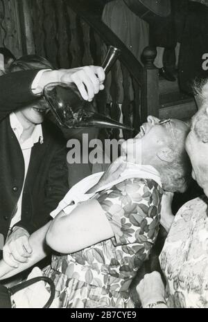 1960 istantanea in bianco e nero di una signora anziana che indossa una rana fiorita che viene forzato vino o sangria da un porron da un giovane, mentre un'altra signora anziana guarda in primo piano. Fotografia vernacolare trovata foto (come immagine Alamy n. 2B7CYE7 senza bordo bianco). Fotografo sconosciuto (collezione Richard Bradley) Foto Stock