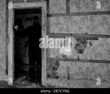 Un uomo che indossa una maschera/cappuccio in un edificio abbandonato. Sta tenendo un coltello grande e sta in piedi nelle ombre in una porta. Bianco e nero. Foto Stock