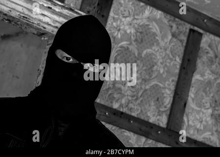 Un uomo che indossa una maschera/cappuccio in un edificio abbandonato. Sta fissando lo spettatore. Vista in primo piano ad angolo. Immagine in bianco e nero scuro. Foto Stock